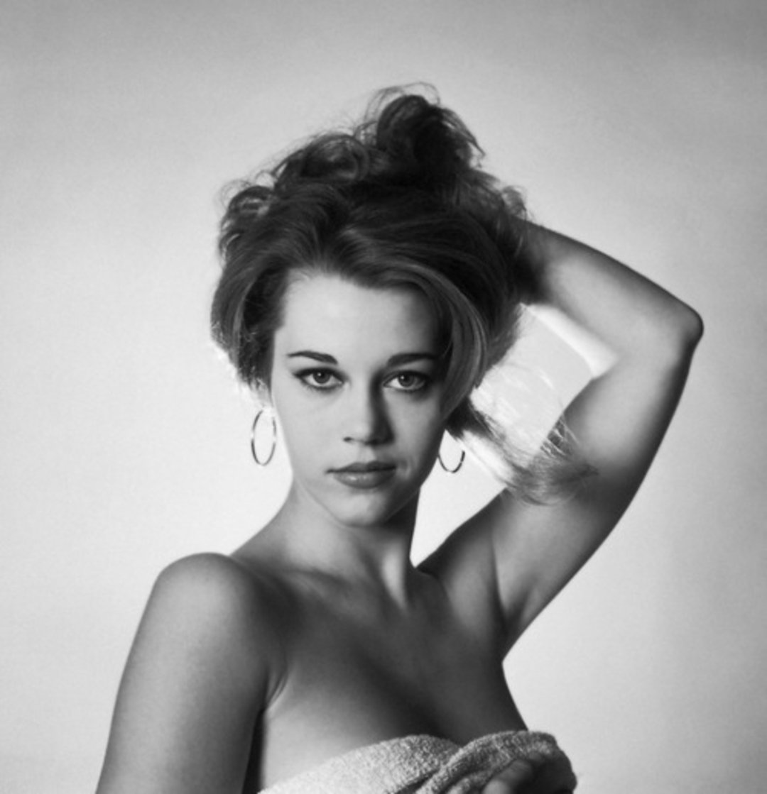 A black and white photo of a woman with voluminous hair, hoop earrings, and dramatic eye makeup. She is wrapped in a towel and poses with one hand lifting her hair, looking directly at the camera.