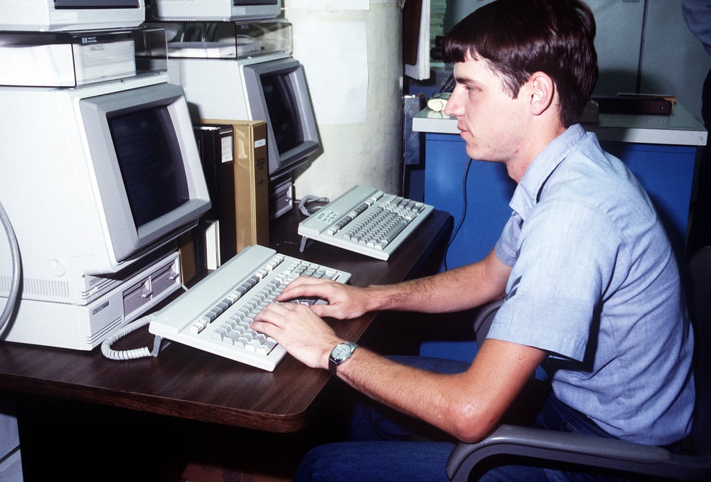 A young man in a short-sleeve shirt types on a vintage computer keyboard at a desk, with two monitors and another keyboard nearby in an office environment.
