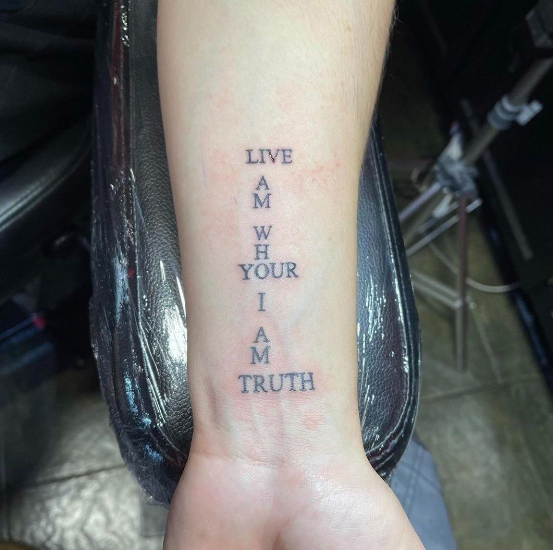 A forearm tattoo with the words "LIVE AM WHO YOUR I AM TRUTH" arranged in a pattern with "AM WHO YOUR I AM" forming a vertical line intersected by "LIVE" and "TRUTH" at the top and bottom.