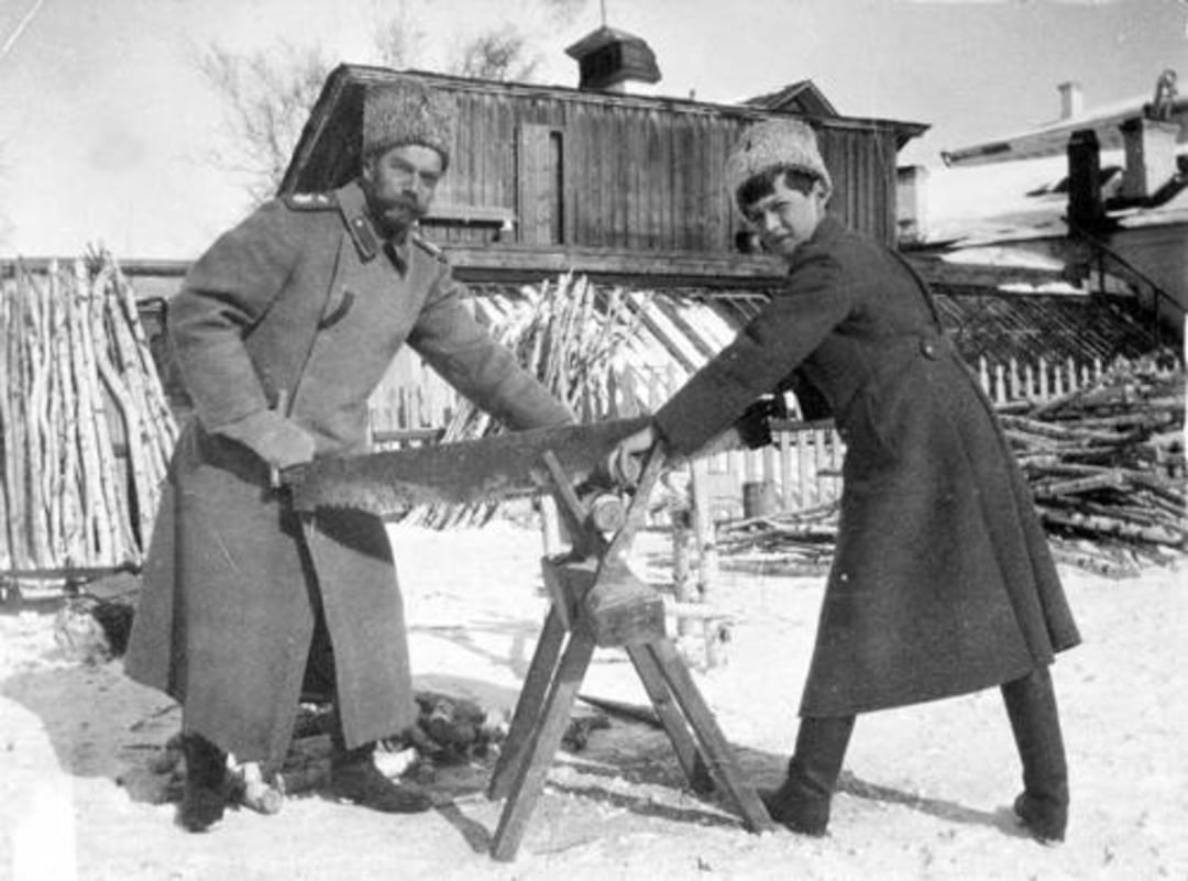 Two men in heavy coats and fur hats use a large handsaw to cut wood outdoors in a snowy area, with a wooden building and stacked logs in the background.
