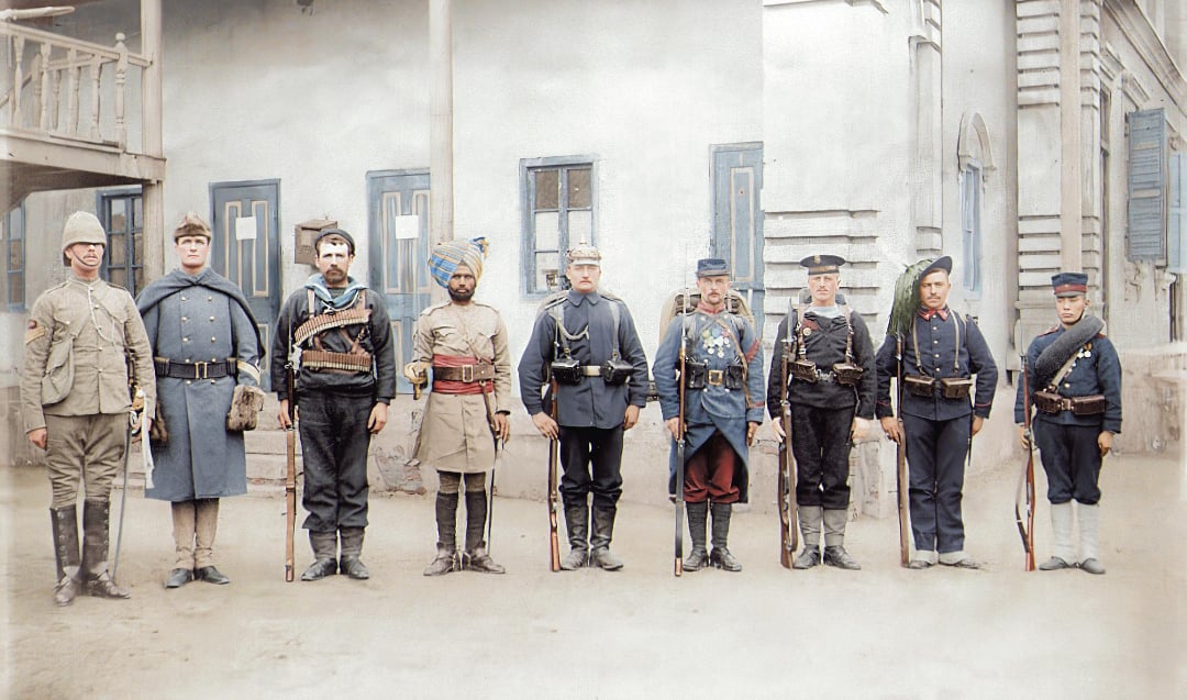 Nine soldiers from different nations in early 20th-century military uniforms stand in a row with rifles, in front of a light-colored building with blue shutters and wooden details.