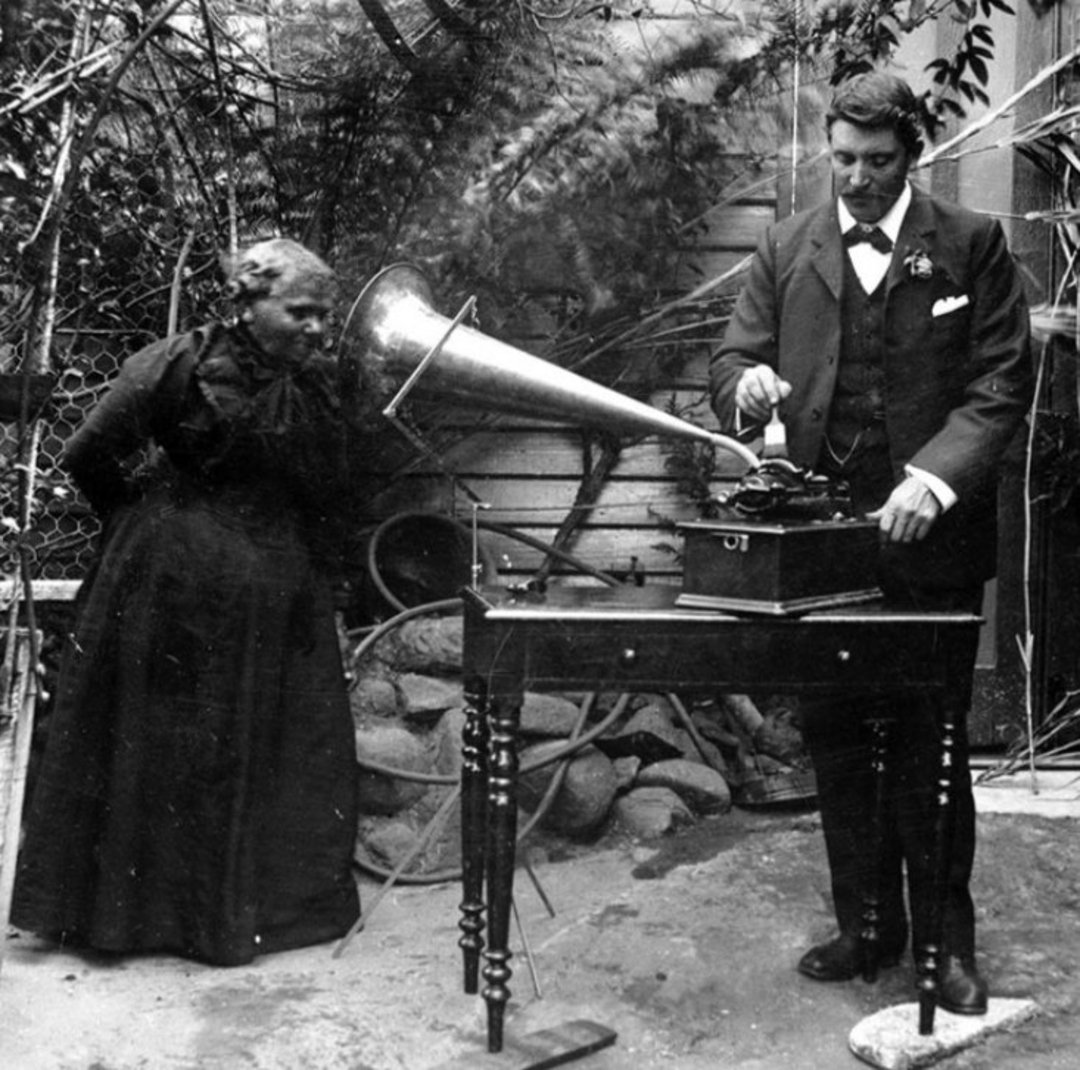 An elderly woman listens to a large horn gramophone while a man in a suit operates it outdoors. The scene is set in a garden with plants and rocks in the background.