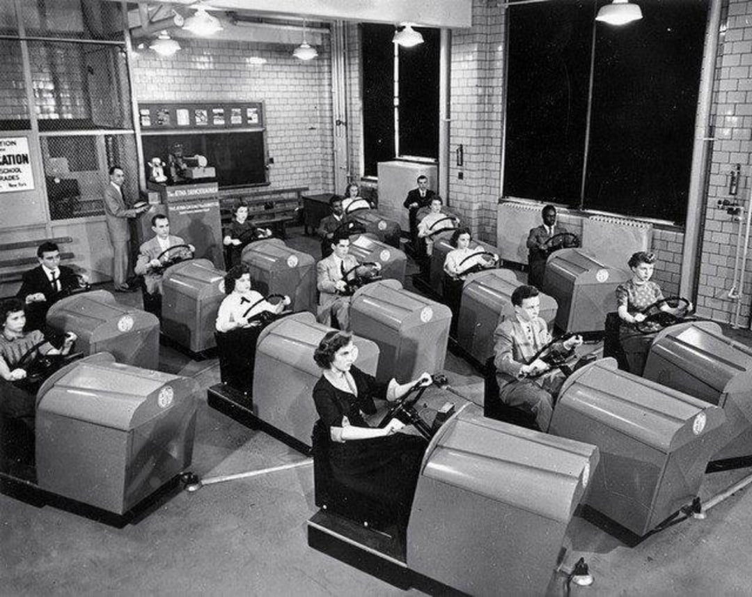 Black and white photo of people sitting in rows inside driving simulators, holding steering wheels, in a classroom setting with an instructor at the front near a chalkboard.