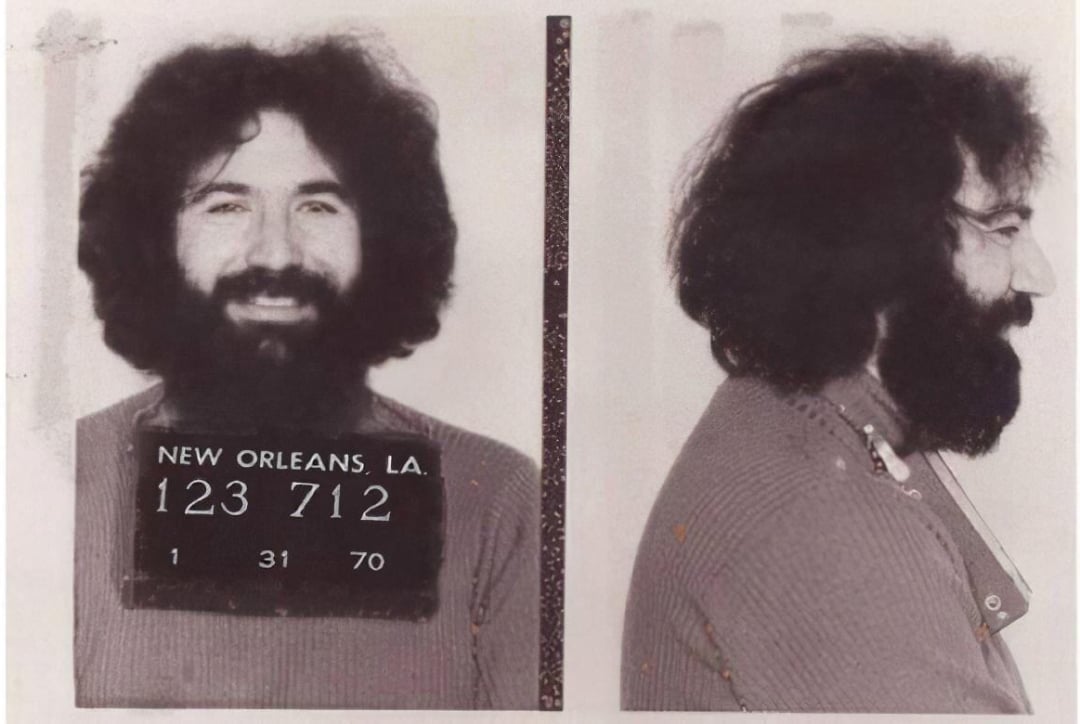 Black-and-white mugshot of a bearded man with curly hair, smiling in the front view. His clothing appears casual. The sign reads "NEW ORLEANS, LA. 123 712 1 31 70." Side profile is also shown.