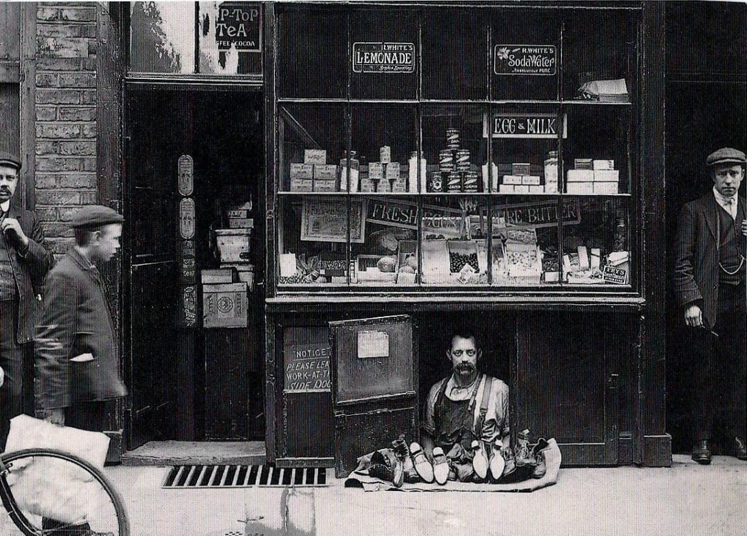 A black-and-white photo of an old shop with shelves of goods and signs in the window. A man sits in a hatch below the shop window, selling shoes, while three other men stand or walk nearby on the street.