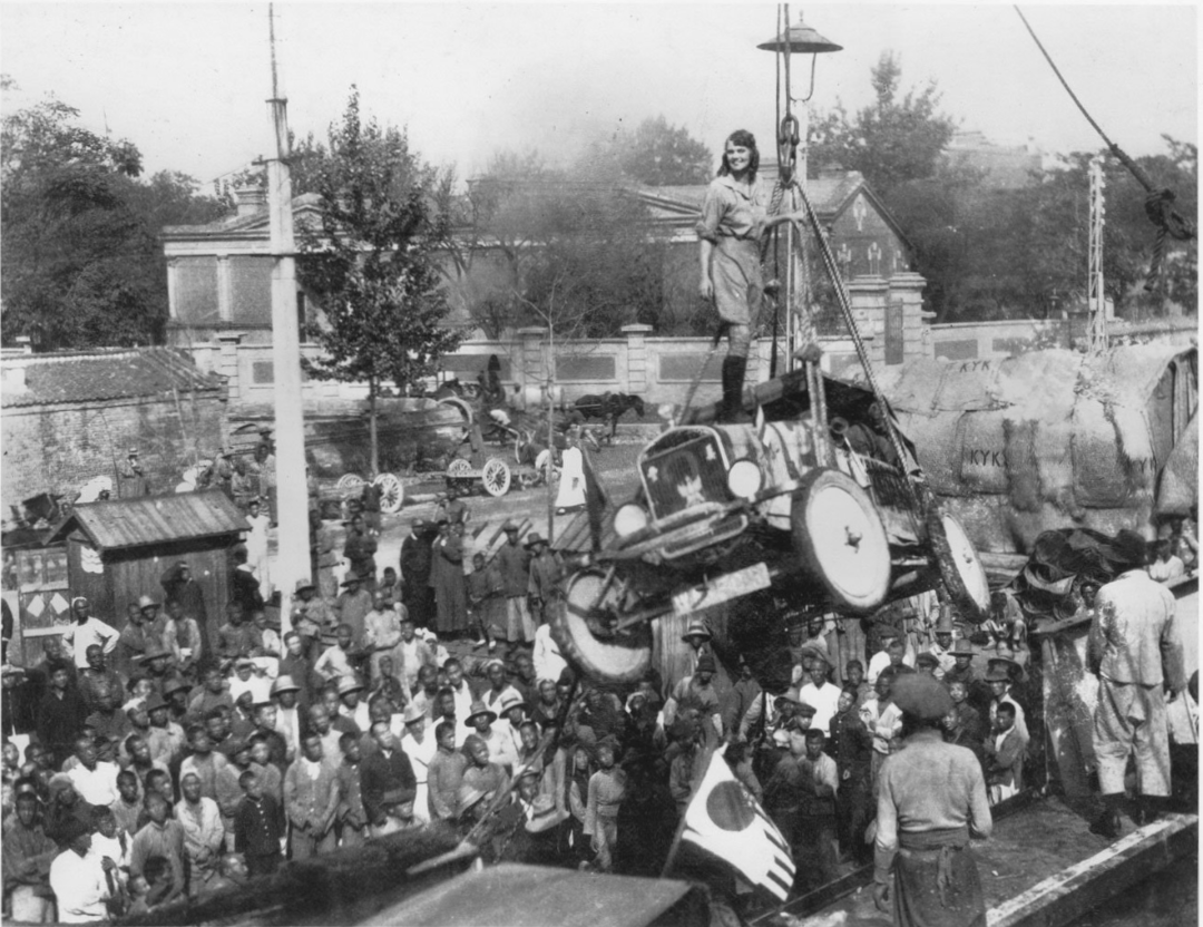 A crowd gathers around as a woman stands on a car being hoisted by a crane above a busy street. People look on from below, with buildings and trees visible in the background. The scene appears historical.