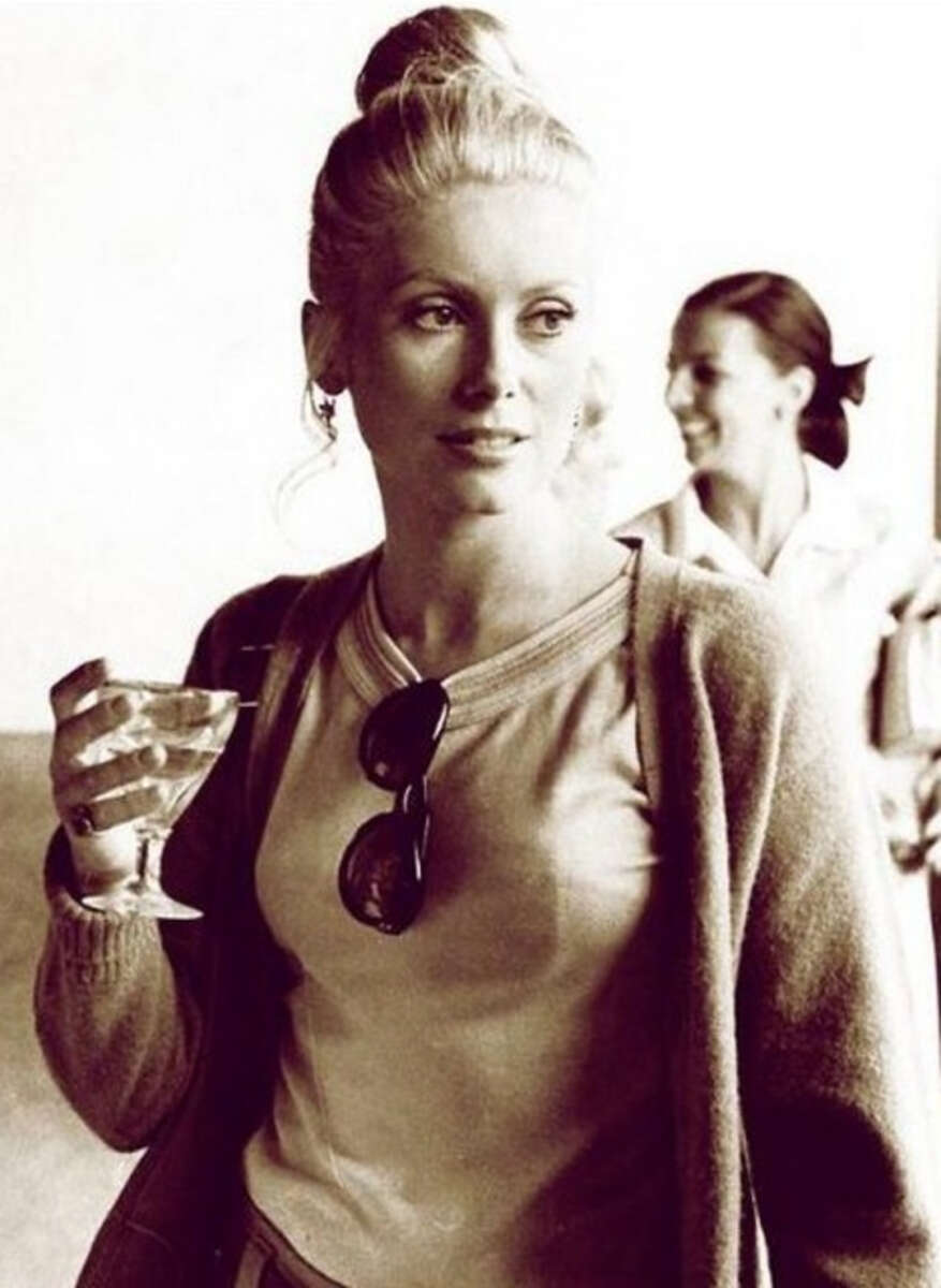 A woman with blonde hair in a bun holds a drink in her right hand and wears a cardigan over a t-shirt with sunglasses hanging from the neckline. Another woman stands blurred in the background, both appear to be indoors.