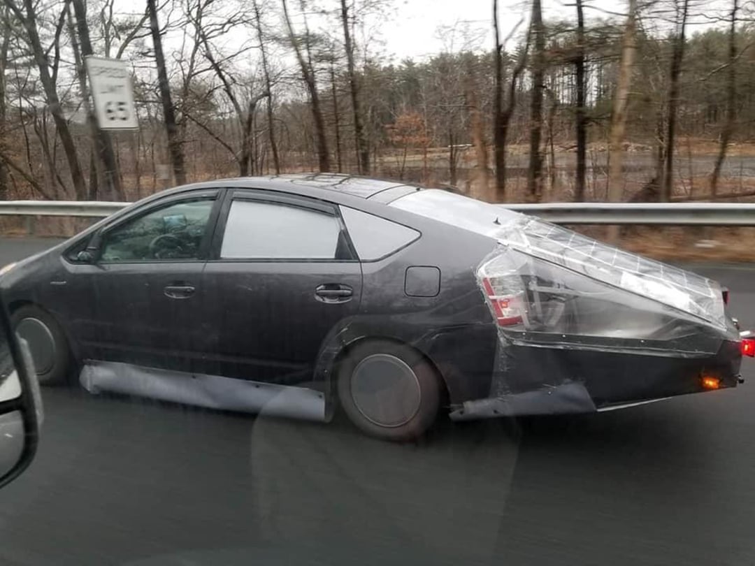 A black car with a large, makeshift, transparent extension taped to its rear drives on a highway. The extension appears homemade and aerodynamic. Trees and a 65 mph speed limit sign are visible in the background.