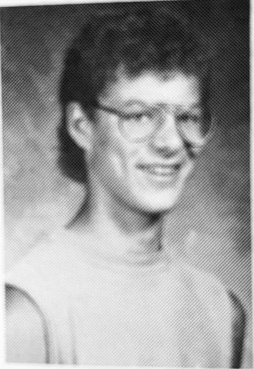 A black-and-white, slightly grainy yearbook-style portrait of a young person with curly hair, glasses, and a sleeveless shirt, smiling at the camera.