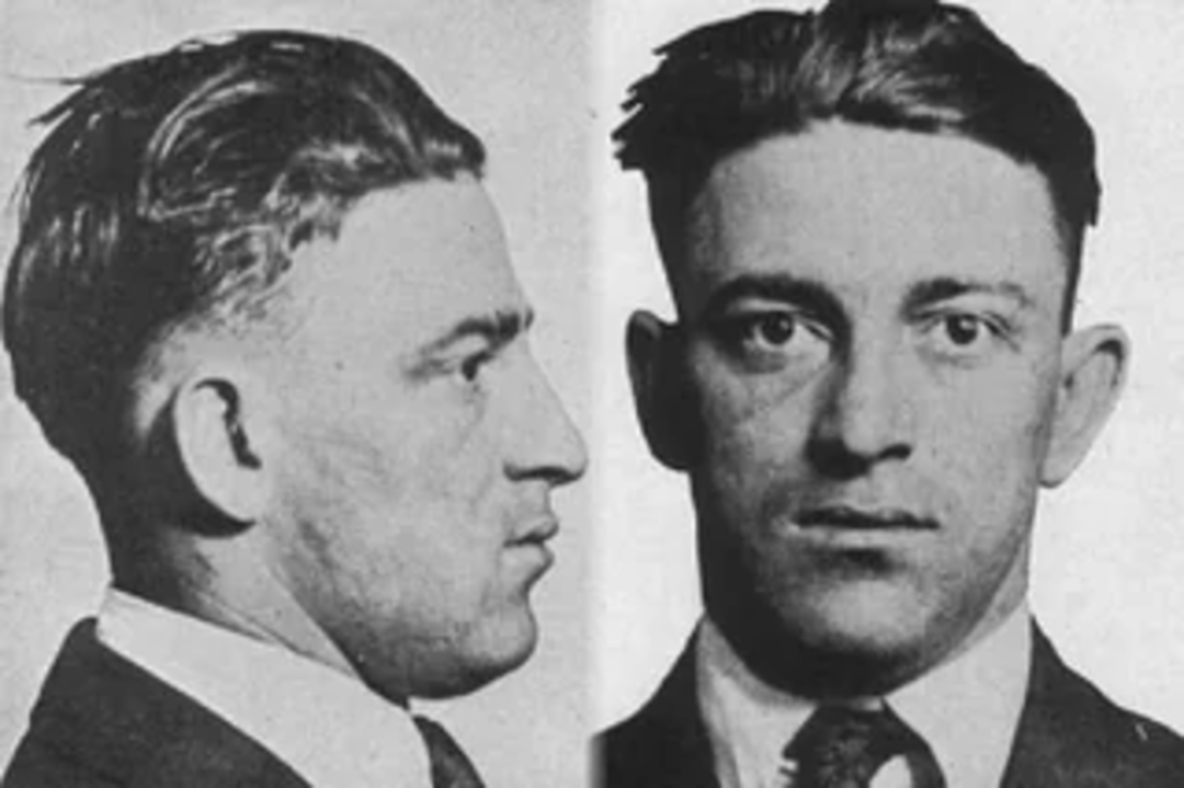 Black-and-white mugshot showing a man with short, dark hair in two views: side profile on the left and front-facing on the right. He wears a suit jacket, dress shirt, and patterned tie.