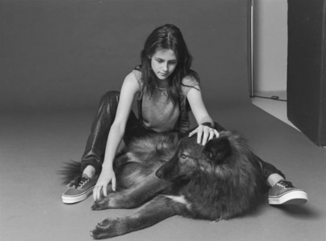 A young woman sits on the floor in a studio, legs crossed, gently petting a large, dark-furred dog lying beside her. The photo is in black and white, with a plain background.