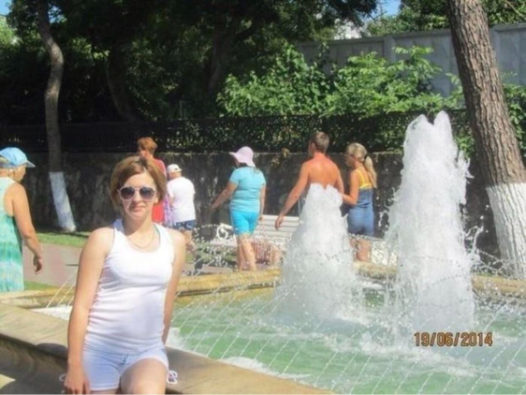 A woman in white clothes and sunglasses poses by a fountain in a park, while several people walk and stand nearby on a sunny day. Trees and greenery are in the background. The date stamp reads 19/06/2014.