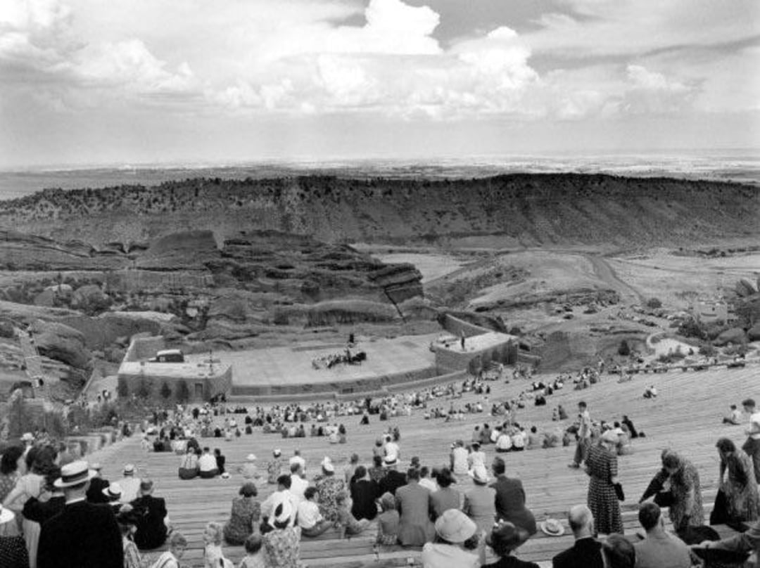 Black and white photo of people sitting on tiered benches at an outdoor amphitheater surrounded by rocky hills, watching a performance on a stage; wide open landscape in the background.