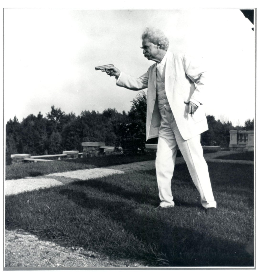 An older man with white hair and a mustache, dressed in a light-colored suit, stands outdoors on grass and points a pistol with one hand while holding his hat in the other. Trees and headstones are visible in the background.