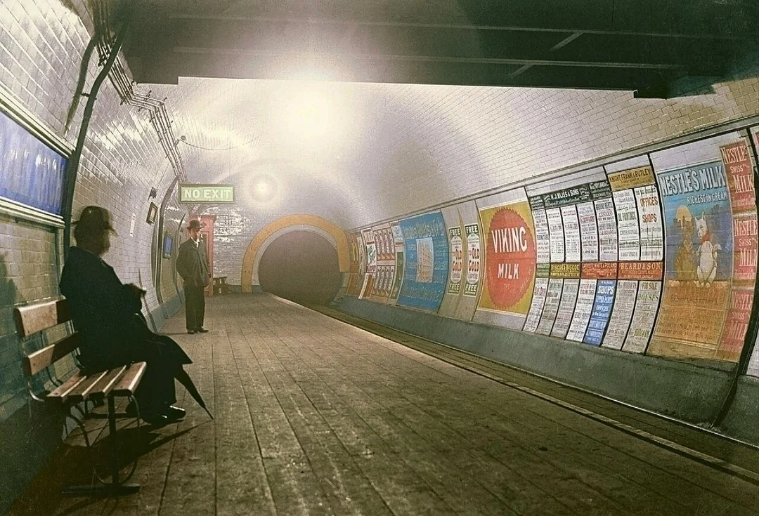 A vintage subway tunnel with wooden floors, two people waiting—one sitting on a bench and another standing. The tunnel walls are lined with colorful, old-fashioned advertisements. A "No Exit" sign is visible.