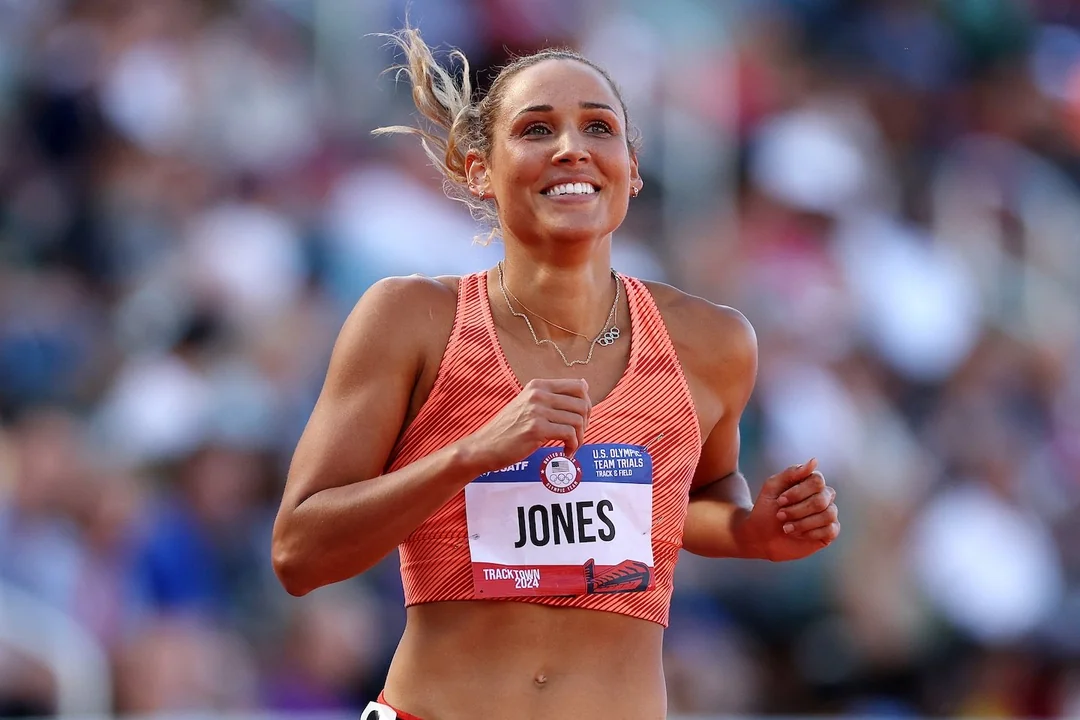 A female athlete wearing an orange sports top and a race bib labeled "Jones" smiles while running on a track, with a blurred crowd in the background.
