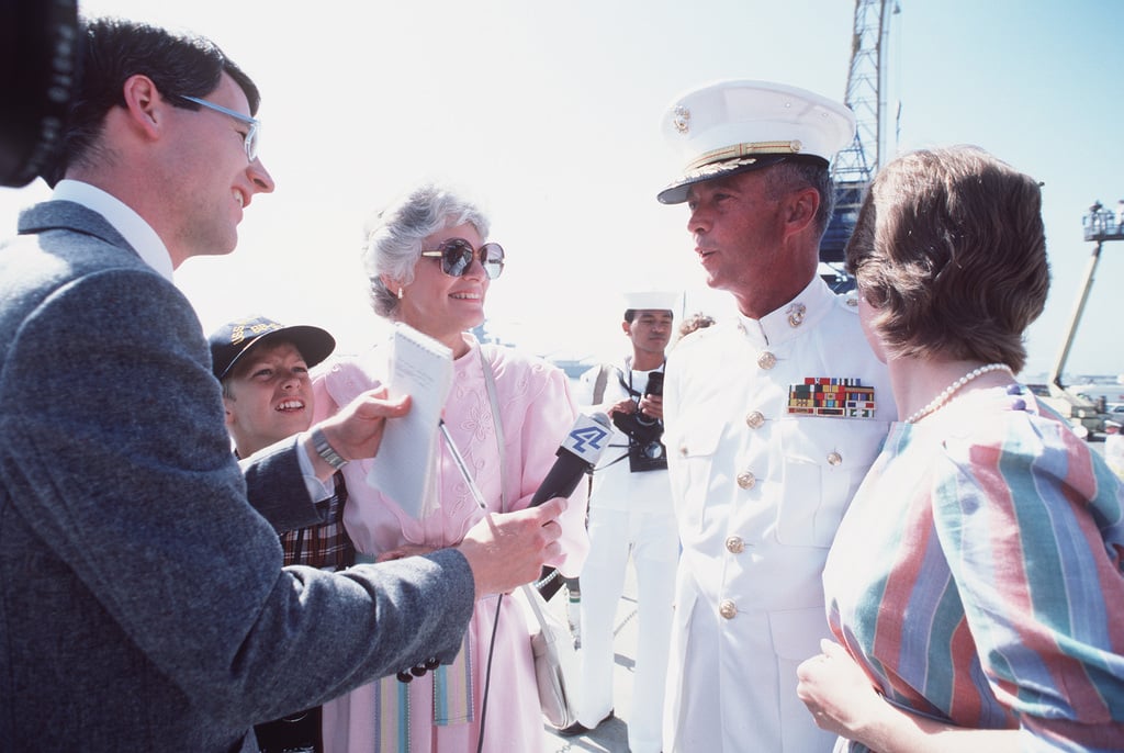 A naval officer in white dress uniform speaks with reporters and two women, one holding a notepad, while a child looks on. Several Navy personnel and industrial equipment are visible in the background.