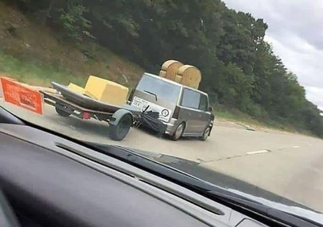 A van drives down a highway towing a trailer with a couch and a canoe, while two large hay bales are strapped to its roof. Trees line the road, and the photo is taken from another vehicle.