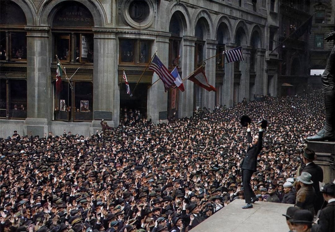 A large crowd of people in hats fills the street outside a grand building with arched windows, listening to a man standing on a platform waving his hat; various flags, including American and French, hang overhead.