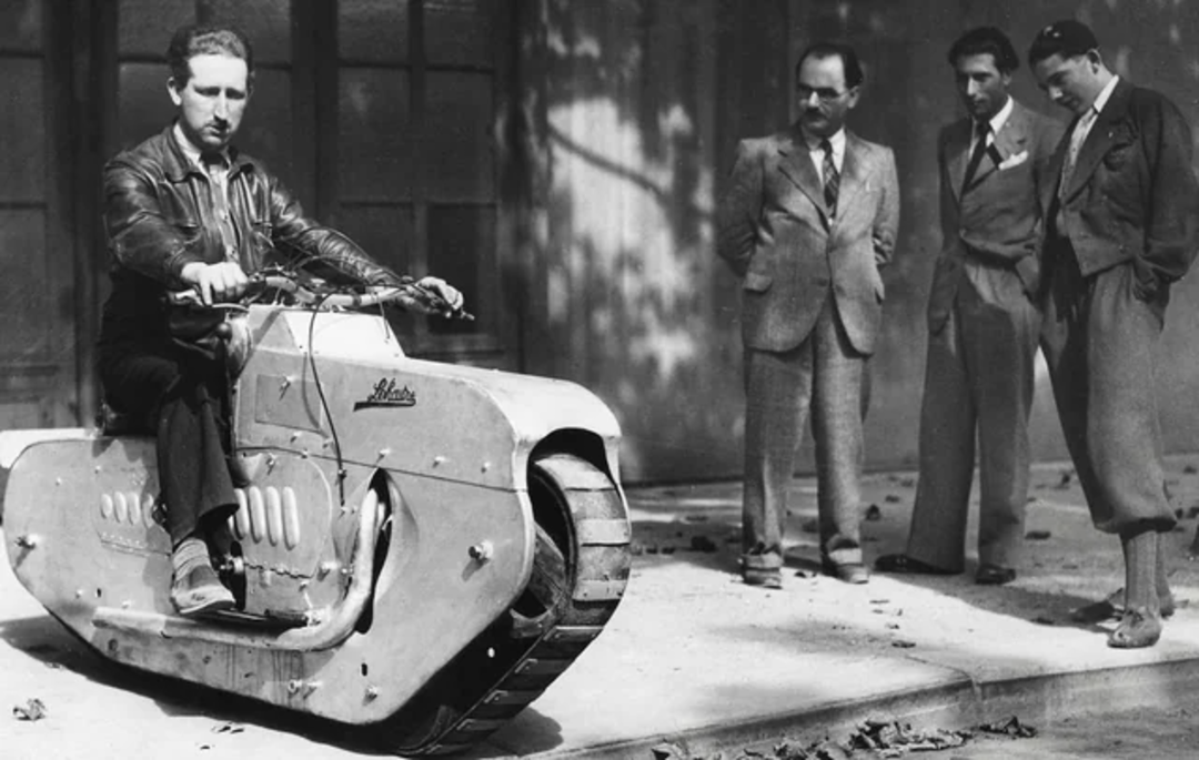 A man sits on a vintage, tank-like motorcycle called a “motorwheel,” while three men in suits stand nearby, watching with interest. The scene appears to be from the early to mid-20th century.