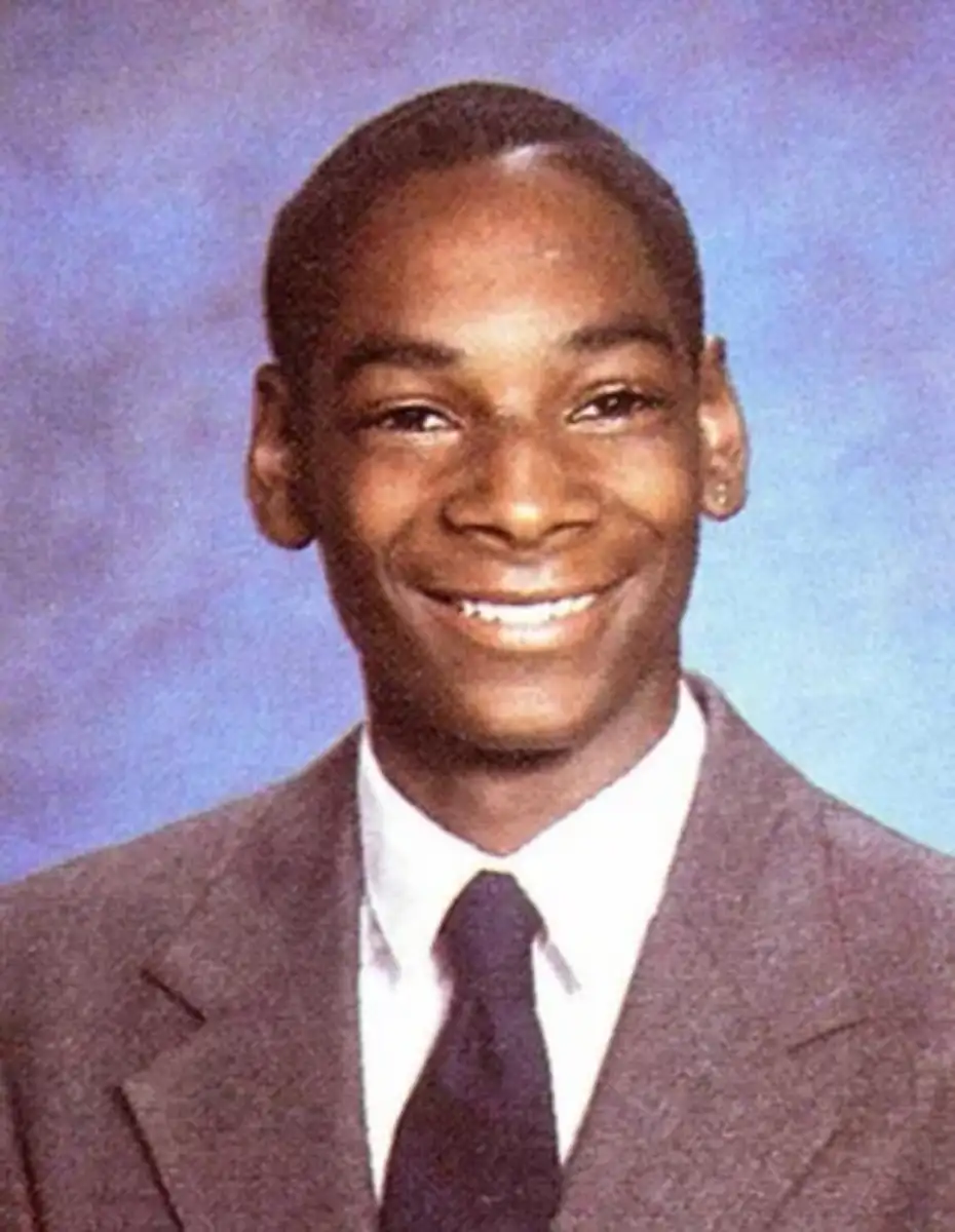 A young man in a brown suit, white shirt, and dark tie smiles in a formal portrait against a blue background, similar to a school yearbook photo.