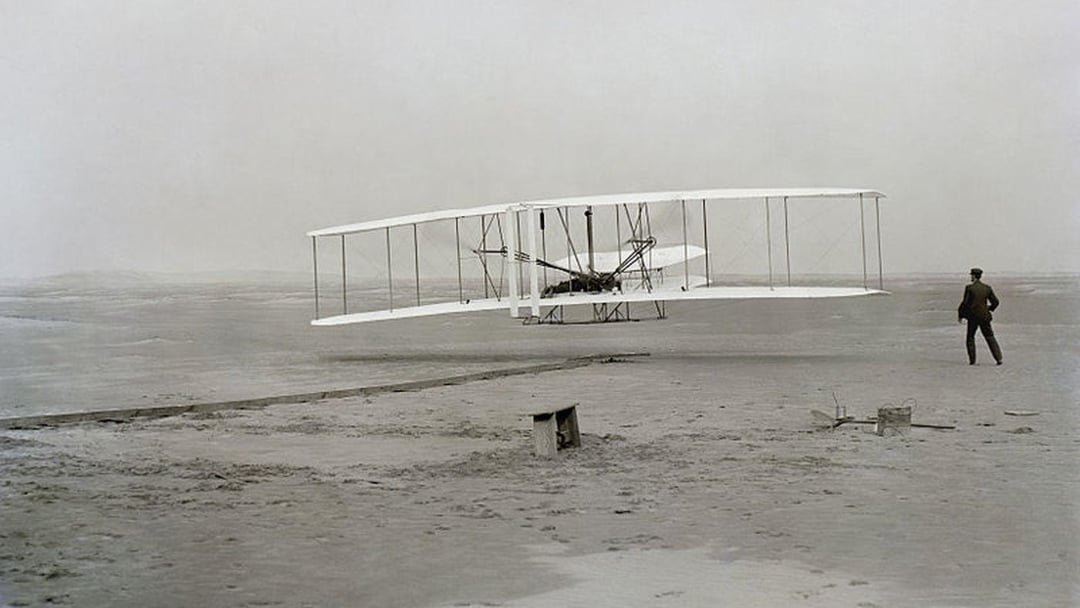 A black and white photo of the Wright brothers’ early biplane flying just above the sandy ground, with one person watching nearby. The landscape is flat and barren, with minimal structures in the background.