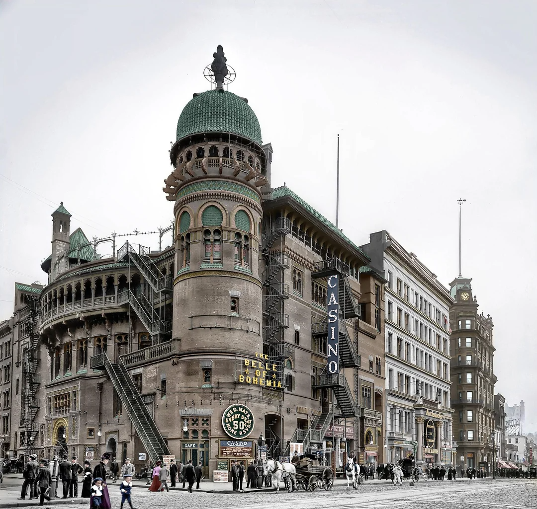 Historic street scene featuring the ornate Casino Theatre with a domed tower, signs reading "Casino" and "Belle of Bohemia," horse-drawn carriages, and people walking on a busy city sidewalk.