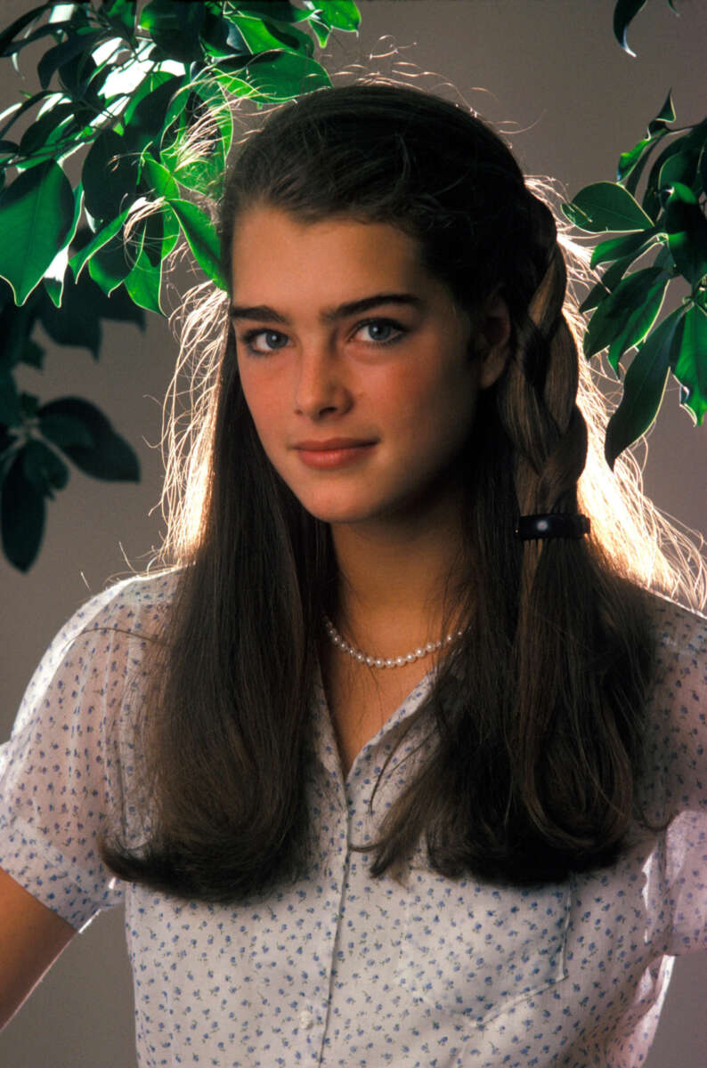 A young woman with long brown hair in a braid, wearing a white patterned blouse and a pearl necklace, stands in front of leafy green branches with sunlight shining behind her.