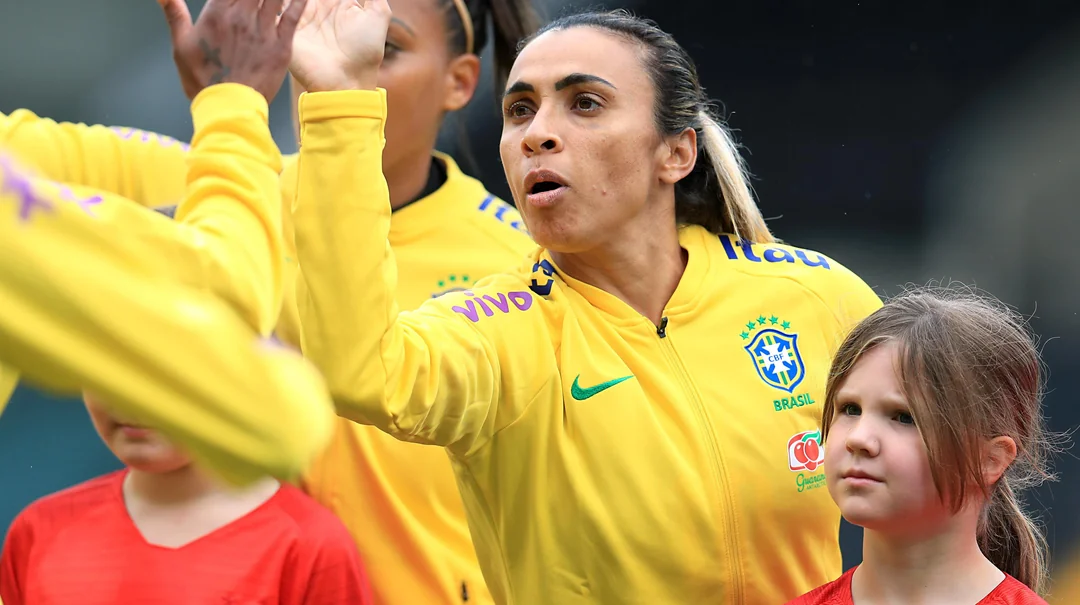 A Brazilian soccer player in a yellow jacket high-fives a teammate, while a young girl in a red shirt stands nearby looking forward. Other team members are partially visible in the background.