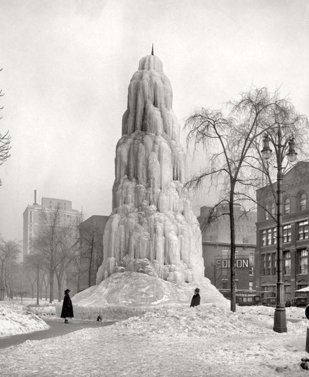 A towering ice sculpture resembling a frozen fountain stands in a snowy city park, with people in winter clothing walking nearby and buildings visible in the background.