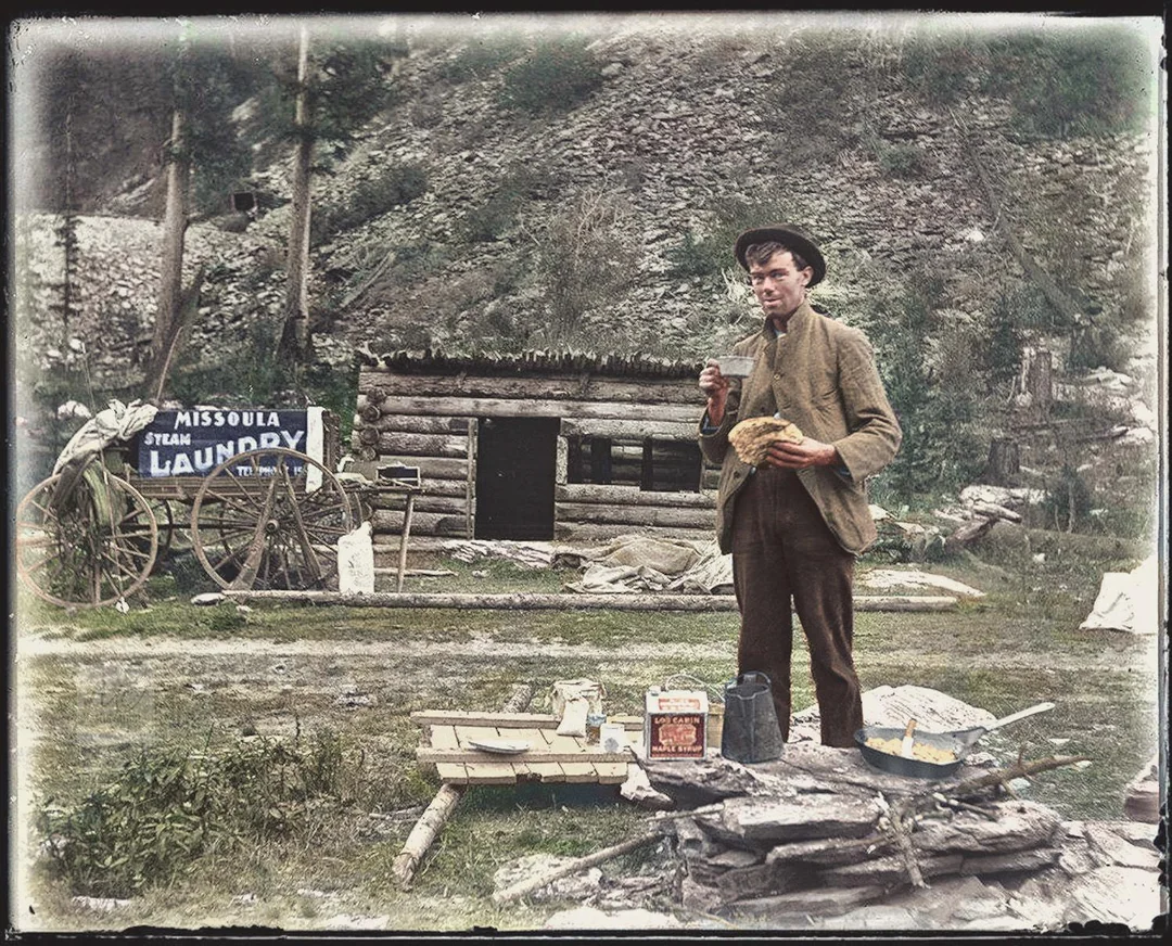 A man in old-fashioned clothes stands outdoors eating food near a log cabin with a "Missoula Steam Laundry" sign. Camping supplies and pans are scattered on the ground around him, with a wooded hillside in the background.
