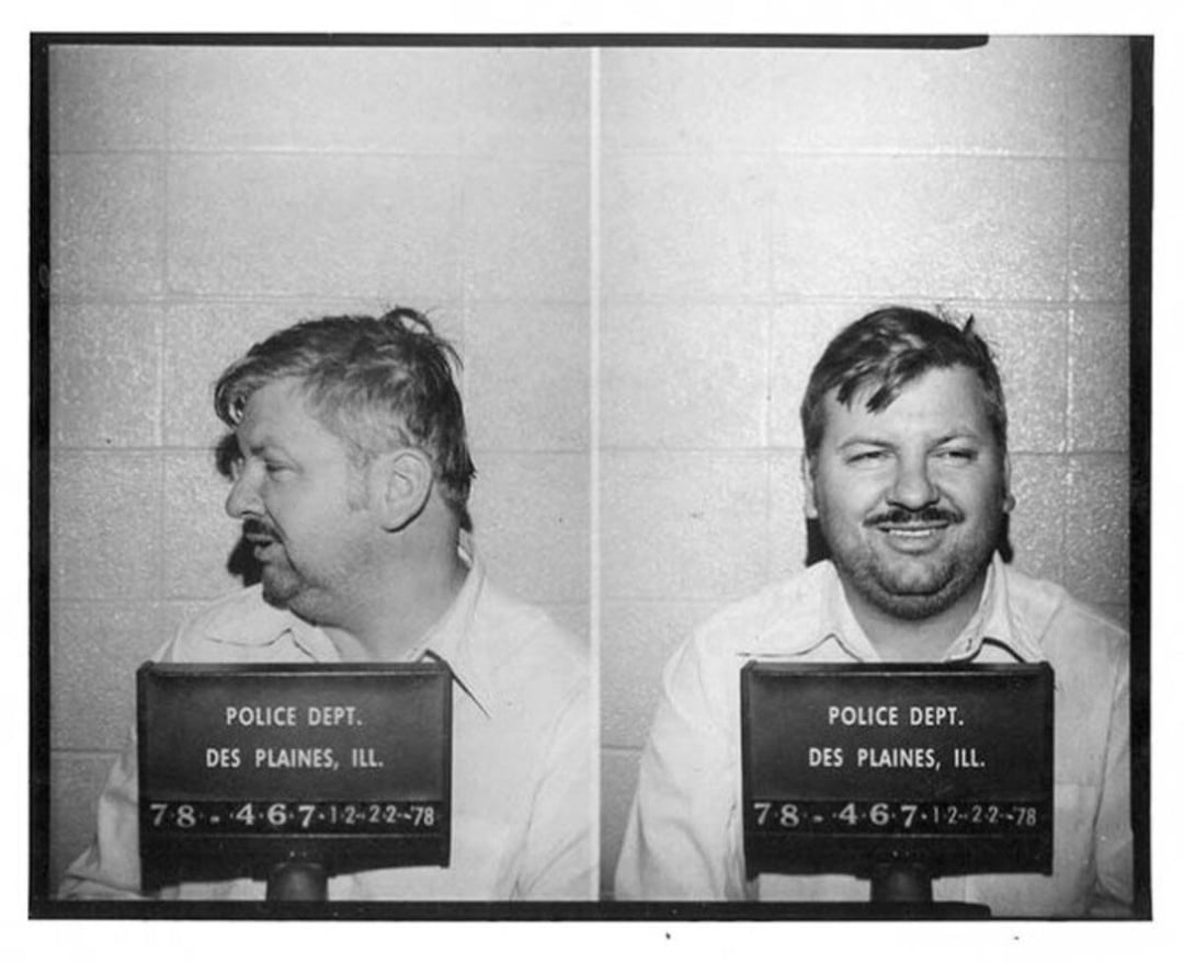 Black and white mugshot of a man with short hair and a mustache, photographed from the side and front, holding a sign reading "Police Dept. Des Plaines, Ill." against a brick wall background.