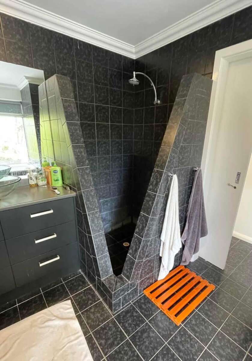 A black-tiled bathroom features a unique, V-shaped walk-in shower with two towels hanging on the side and a wooden slatted mat in front of the shower entrance. A vanity with a sink and mirror is nearby.