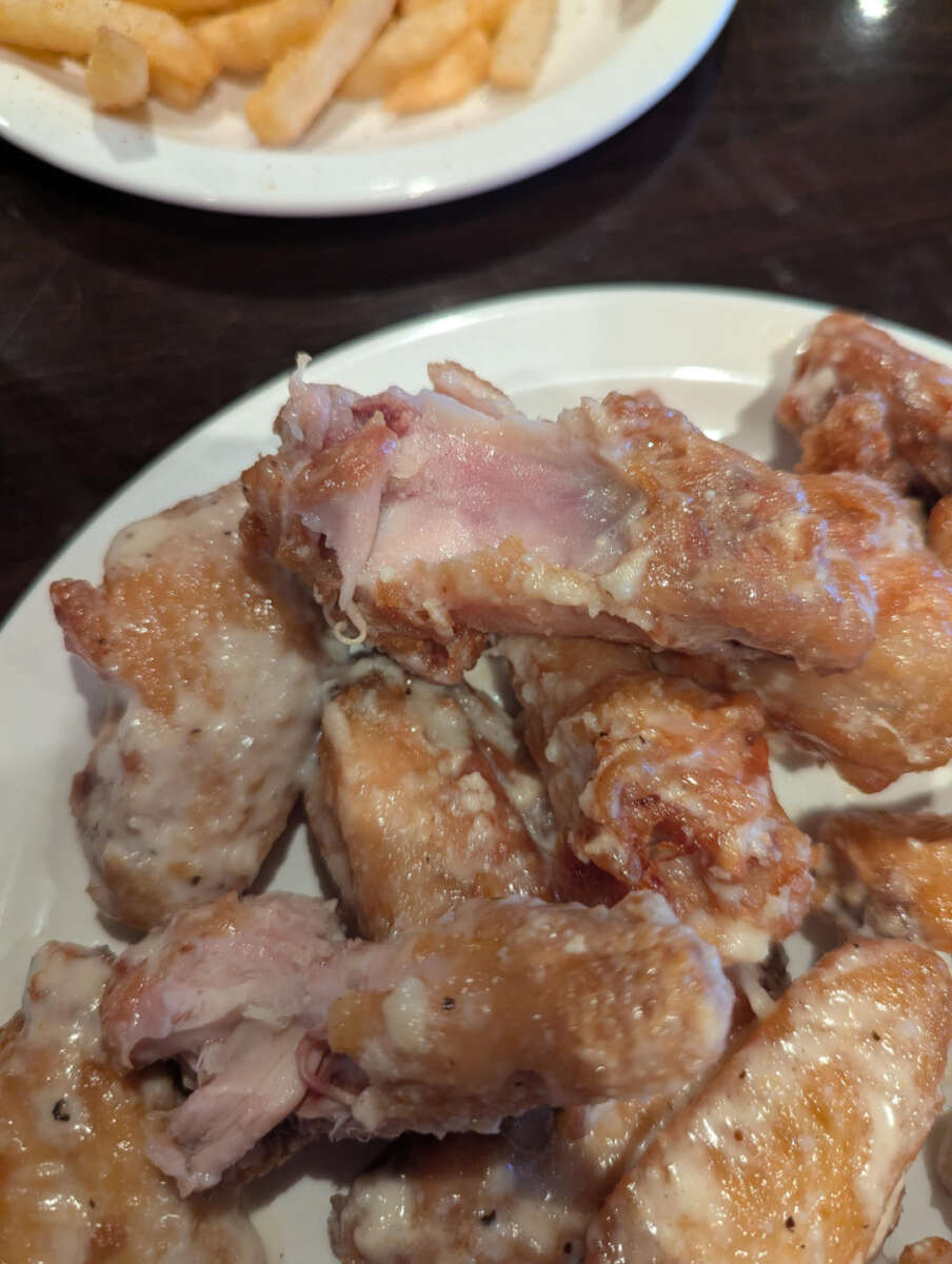 A plate of chicken wings with pink, undercooked meat is shown in the foreground. In the background, there is a plate of crinkle-cut French fries.