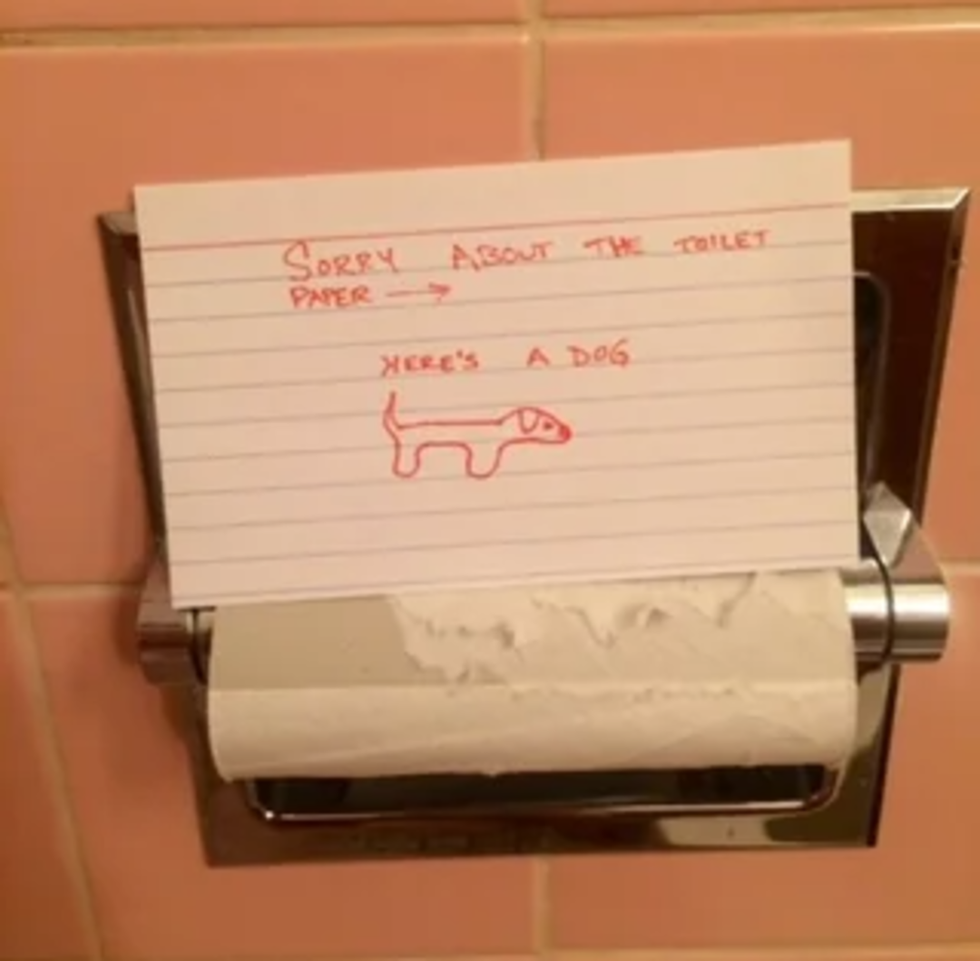 An empty toilet paper holder with a handwritten note that says, "Sorry about the toilet paper," and a drawing of a dog labeled "Here’s a dog." The holder is mounted on a pink tiled wall.