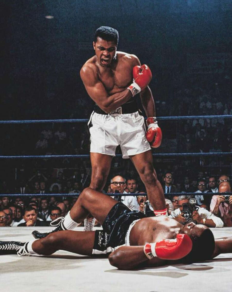 A boxer in white trunks and red gloves stands over his fallen opponent in black trunks, shouting, while a crowd watches from ringside in a dramatic boxing match moment.