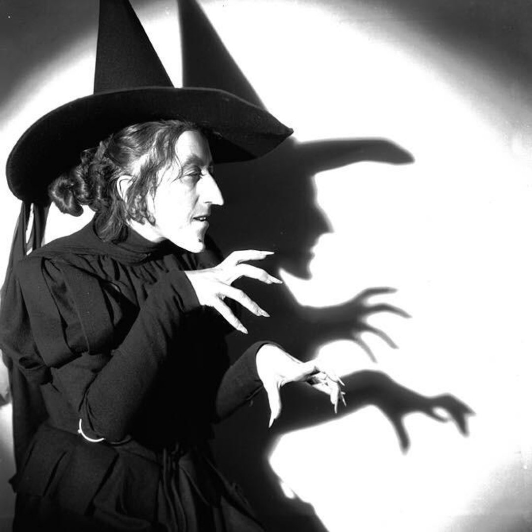 A person dressed as a classic witch with a pointed hat and long, claw-like fingers poses dramatically; their shadow mimics the pose on the wall behind them. The image is in black and white.