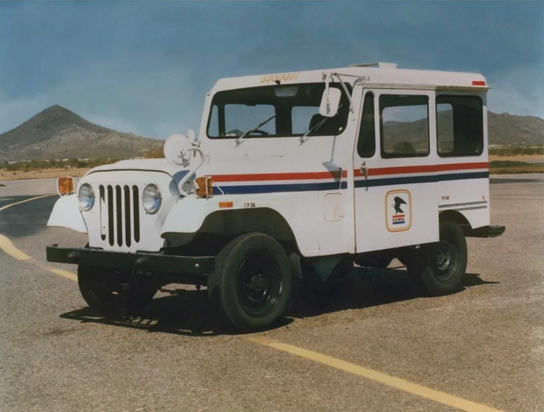 A vintage white postal delivery jeep with red and blue stripes and a U.S. Mail logo is parked on an empty road with mountains visible in the background under a clear sky.