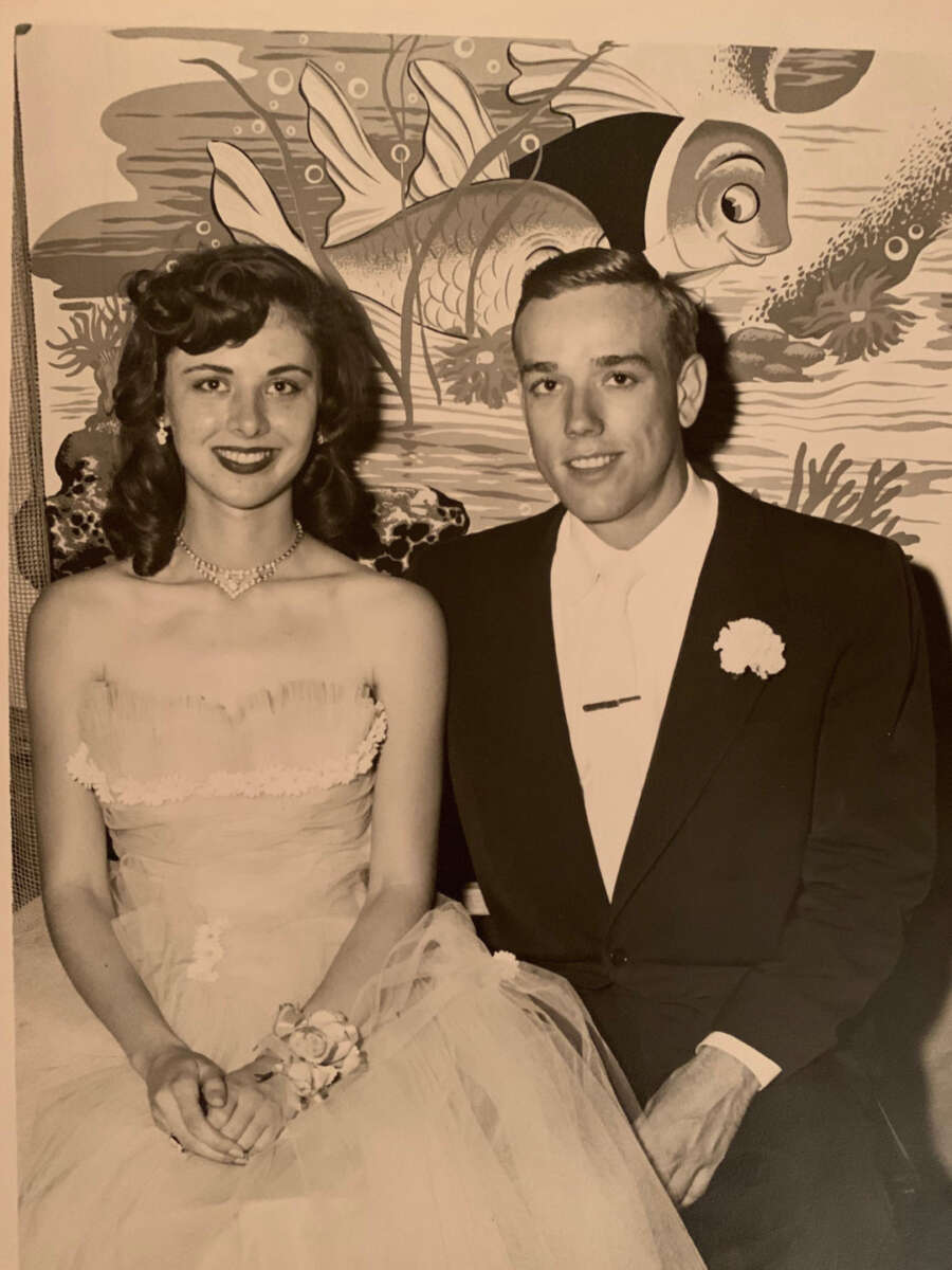 A young woman in a strapless dress and corsage sits next to a young man in a suit with a boutonniere. They are smiling and seated in front of a colorful fish-themed mural.