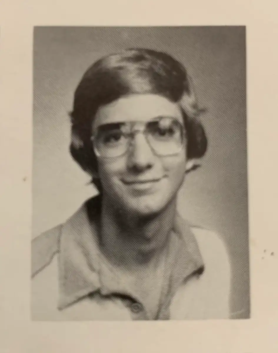 A black-and-white yearbook photo of a young person with medium-length hair, wearing large glasses and a collared shirt, smiling slightly at the camera.