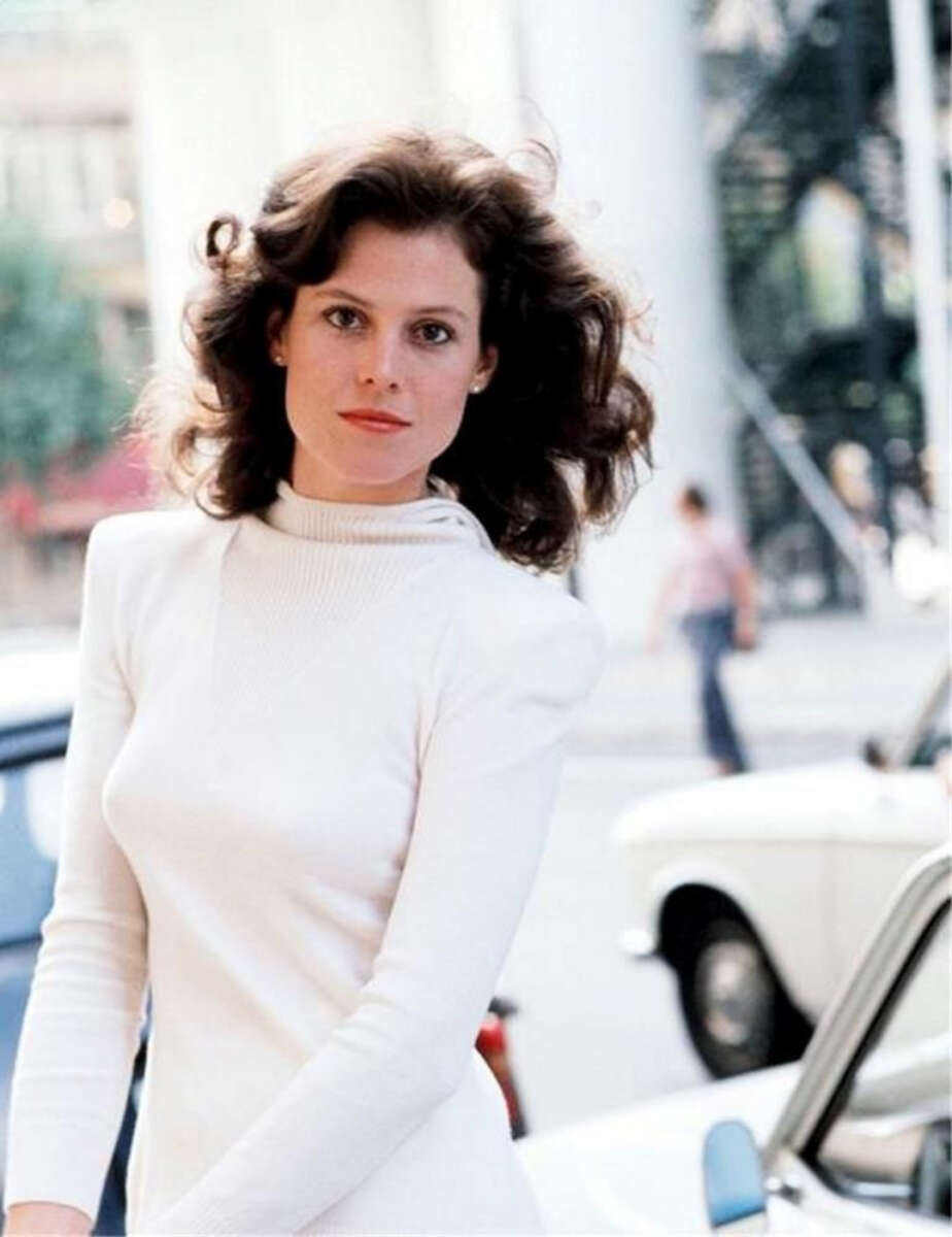 A woman with wavy brown hair wearing a long-sleeved white turtleneck dress stands outdoors near a white car, with blurred street and buildings in the background. She looks directly at the camera with a calm expression.