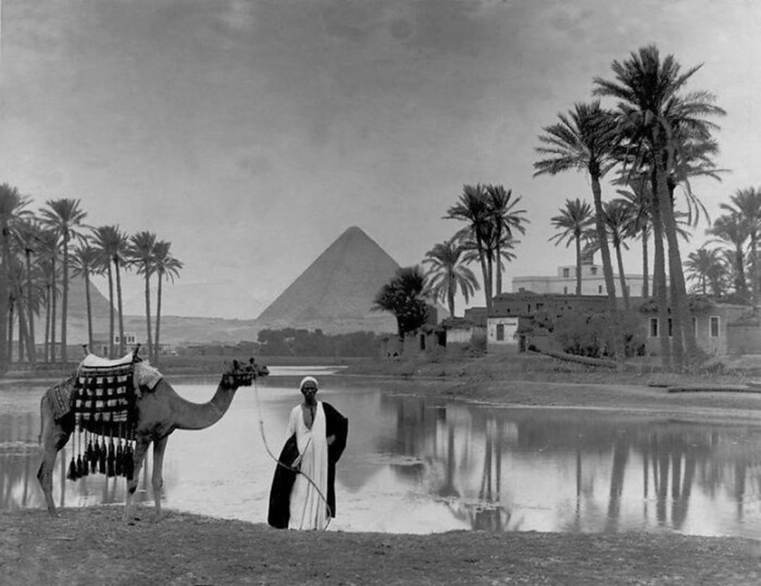 A man in traditional clothing stands beside a camel near a reflective body of water, with palm trees, houses, and the Great Pyramid of Giza visible in the background.