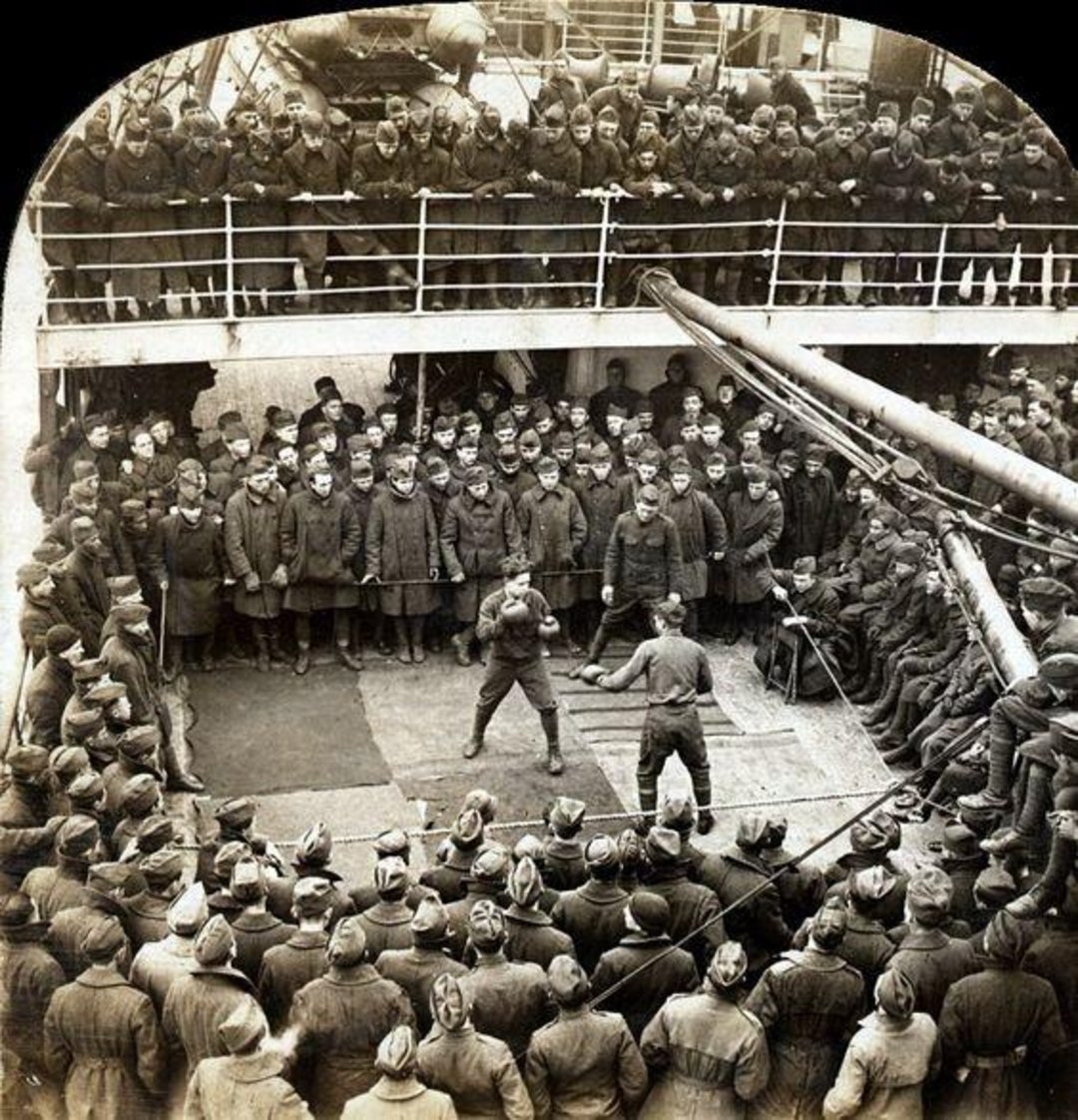 A large crowd of soldiers on a ship watches two men boxing at the center of the deck. The men are surrounded on all sides, with some spectators standing on an upper level, all wearing military uniforms and caps.
