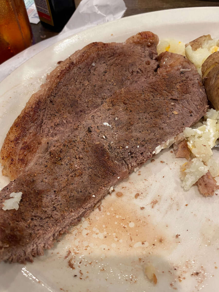 A large, cooked steak seasoned with pepper sits on a white plate next to a baked potato with some of its filling exposed. Some steak juices and potato bits are visible on the plate.