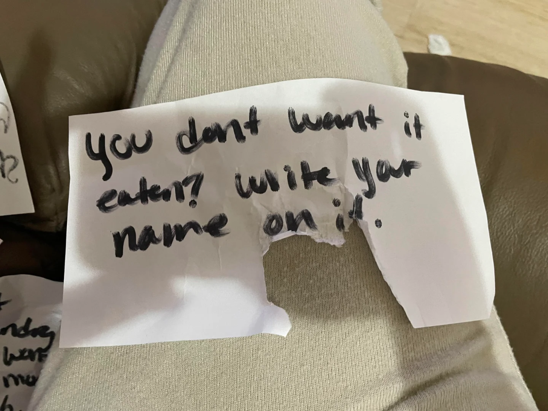 A handwritten note on a torn piece of paper reads, "You don't want it eaten? Write your name on it." The paper rests on someone's khaki-clad lap.