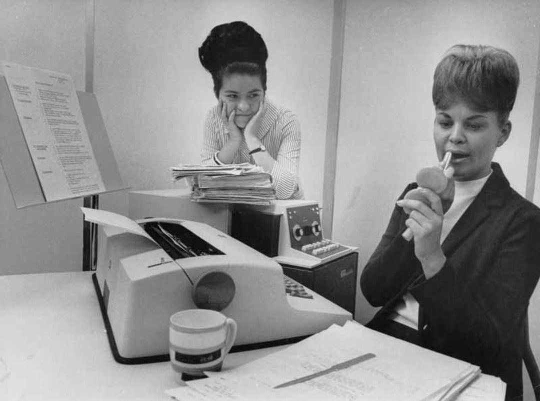 Two women in an office; one sits at a desk with a typewriter, looking in a compact mirror and applying lipstick, while the other woman leans on a stack of papers, smiling and watching her.