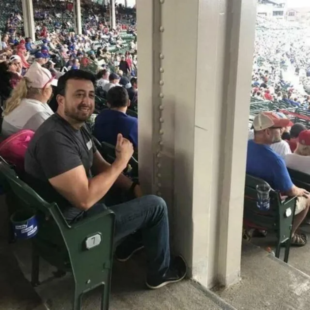 A man sits in seat 7 at a stadium, directly behind a large pillar that blocks his view of the field. He smiles and gestures with his thumb toward the pillar. Many spectators are seated around him.
