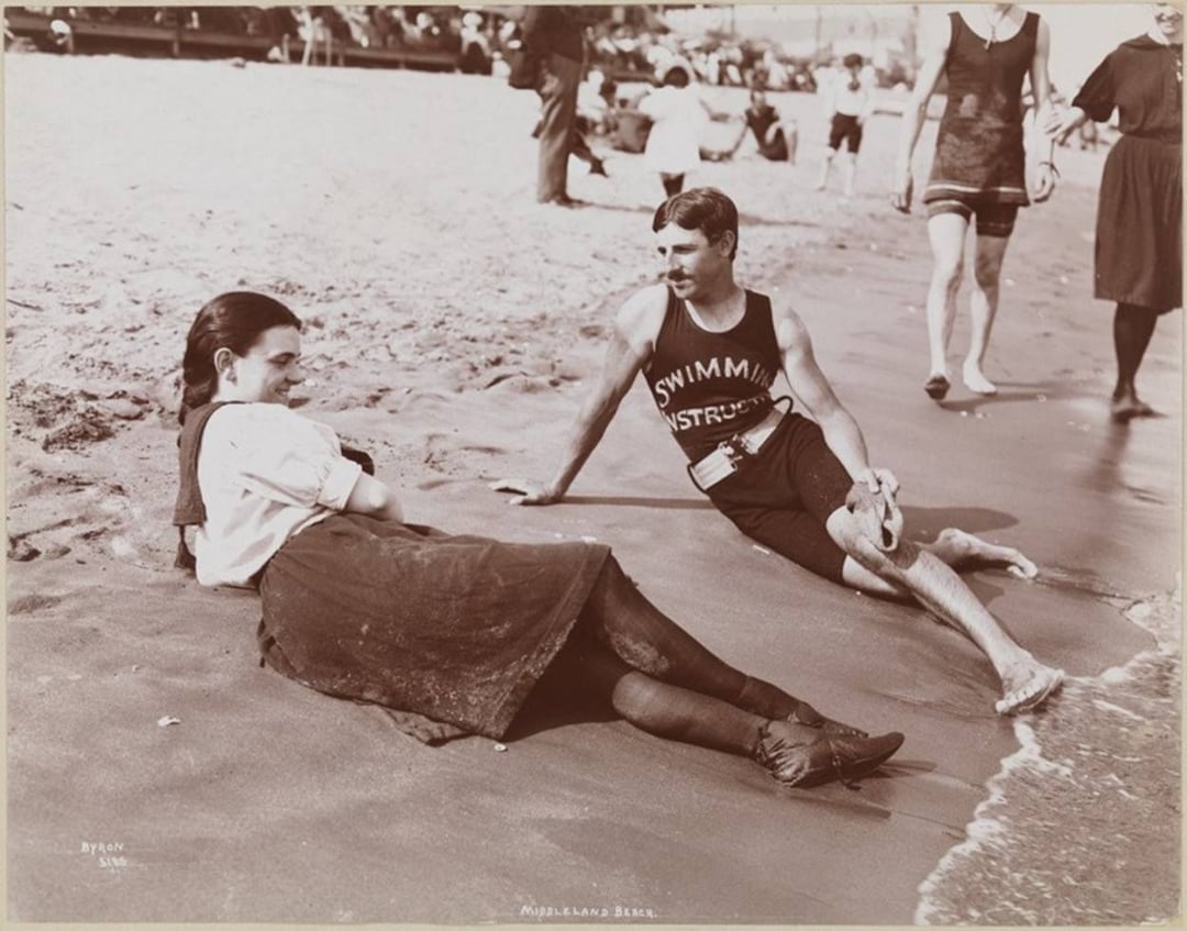 A woman in a dress and a man in a "Swimming Instructor" tank top sit and chat on the sandy shore of a beach, with people and the ocean in the background.