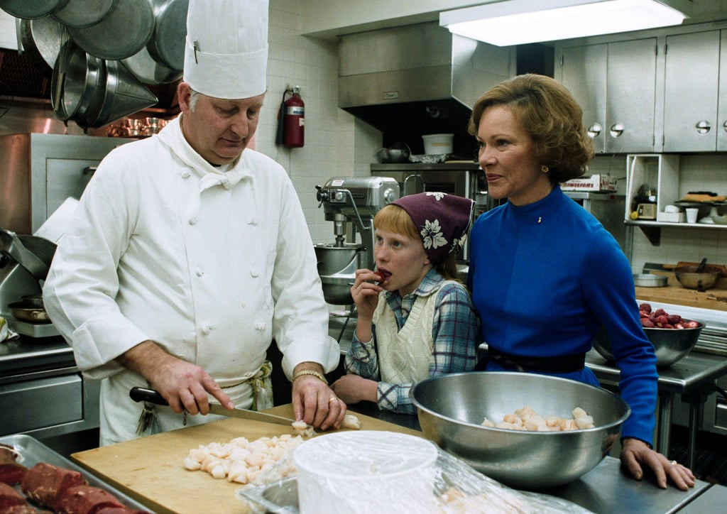 A chef in a white uniform slices food on a cutting board while a young girl and a woman in a blue dress watch in a busy kitchen. The girl snacks on something and the woman stands beside a large metal bowl.