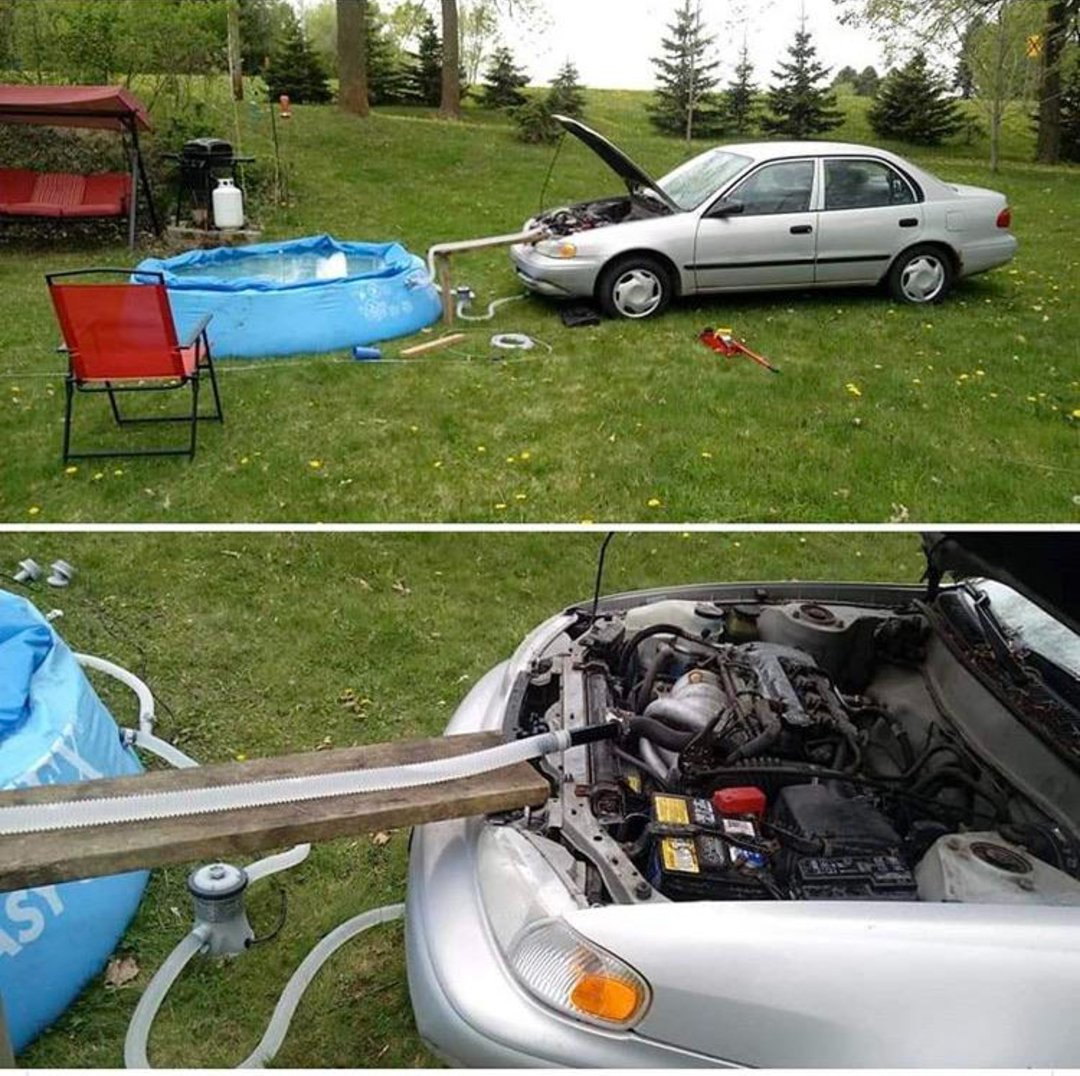 A silver car with its hood open is parked next to a small blue inflatable pool in a yard. Hoses and a metal part connect the car engine to the pool, suggesting it's being used to heat the pool water.